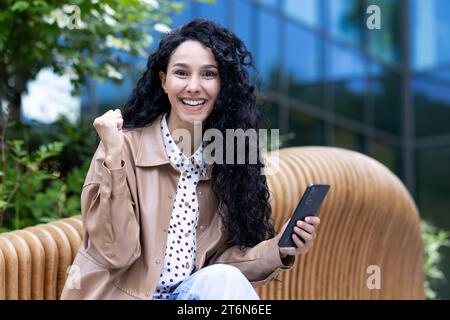 Jeune femme hispanique attrayante avec les cheveux bouclés assis sur le banc à l'extérieur tenant le téléphone dans la main et souriant regardant la caméra, heureuse brunette dans l'excitation de gagner en ligne, obtenir de bonnes nouvelles. Banque D'Images