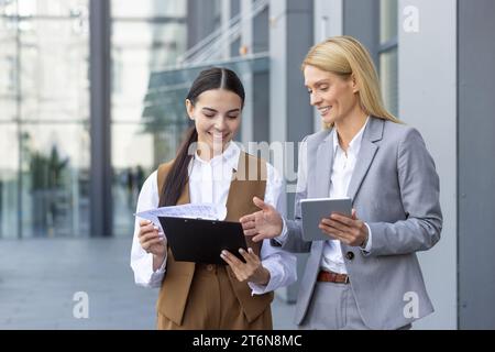Deux collègues d'affaires heureuses en costume debout à l'extérieur du bureau, discutant de questions importantes de travail, comparant des documents, se préparant à une présentation. Banque D'Images