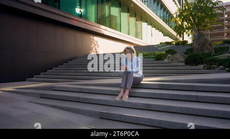 Pleine longueur vue travail d'affaires à l'extérieur caucasien mature senior d'âge moyen femme d'affaires employeur PDG femme aux cheveux gris s'assoient sur les escaliers près du bureau Banque D'Images