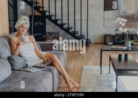 Une femme détendue avec un turban de serviette apprécie le café sur un canapé, avec un magazine Banque D'Images