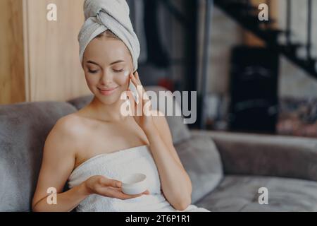 Une femme avec un couvre-tête en serviette applique une crème pour le visage, assise confortablement sur un canapé Banque D'Images