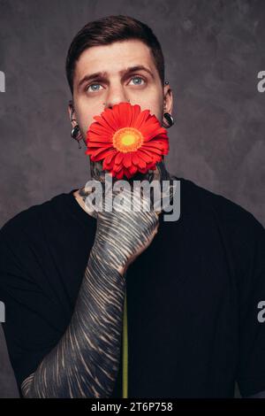 Contemplé jeune homme avec des oreilles de nez percées tenant la fleur de gerbera rouge devant sa bouche sur fond gris Banque D'Images