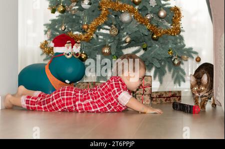 Noël. Un petit garçon beau en pyjama à carreaux rouges se trouve sur le sol et joue avec une locomotive à vapeur rouge et un chat Bengal contre le Banque D'Images