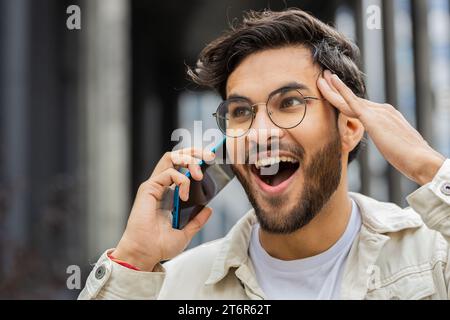 Appel téléphonique, bonnes nouvelles, commérages. Heureux surpris homme indien dans une conversation agréable sur smartphone, profiter de parler avec un ami à l'extérieur. Arabian excité gars debout dans la rue de la ville. Style de vie urbain Banque D'Images