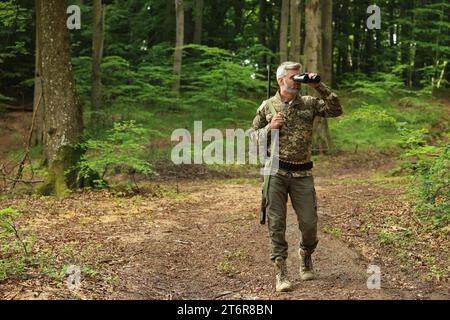 Homme avec un fusil de chasse regardant à travers des jumelles dans la forêt. Espace pour le texte Banque D'Images