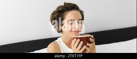 Gros plan portrait de femme sentant son café, prenant la tasse du matin de boisson aromatique préférée, ferme les yeux et souriant, assis sur le lit Banque D'Images