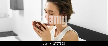 Portrait de jeune femme se réveillant, buvant du café au lit, sentant l'arôme préféré Banque D'Images