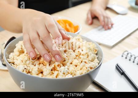 Mauvaises habitudes. Femme mangeant du pop-corn tout en travaillant sur ordinateur à table, gros plan Banque D'Images