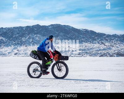 Homme voyageur monte un gros vélo sur un lac de montagne gelé. Voyager en hiver. Expédition extrême. Banque D'Images