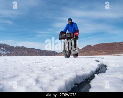 Homme voyageur monte un gros vélo sur un lac de montagne gelé. Voyager en hiver. Expédition extrême. Banque D'Images
