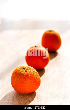 Trois mandarines alignées sur une table en bois devant une fenêtre, la première est mise au point, le reste est flou. Banque D'Images