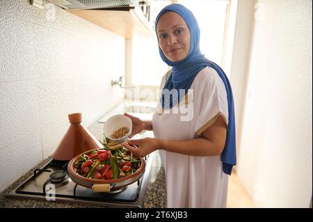 Femme du Moyen-Orient, agréable femme au foyer en hijab et robe authentique, mettant des pois chiches dans un plat avec des légumes, souriant, regardant la caméra wh Banque D'Images