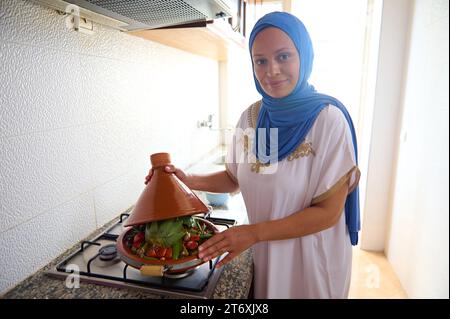 Souriante femme du Moyen-Orient, jolie femme au foyer avec la tête couverte de hijab bleu, debout par le poêle dans la cuisine à la maison, cuisinant délicieux repas marocain Banque D'Images