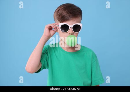 Garçon dans les lunettes de soleil soufflant du bubble gum sur fond bleu clair Banque D'Images