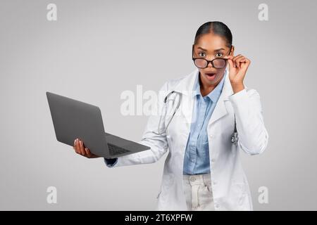 Glad choqué jolie dame noire millénaire médecin en manteau blanc avec ordinateur portable, avec la bouche ouverte, enlever les lunettes Banque D'Images