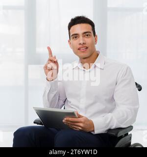 Portrait jeune homme d'affaires assis en fauteuil roulant tenant la main de tablette numérique pointant son doigt vers le haut Banque D'Images