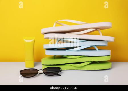 Pile de différentes tongs, lunettes de soleil et crème solaire sur la table blanche sur fond jaune Banque D'Images