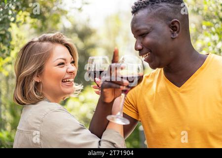 Un couple interracial partage un toast avec des verres de vin rouge, leur rire symbolisant une connexion joyeuse. Banque D'Images