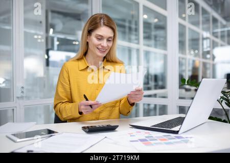 Une femme blonde séduisante travaille dans un bureau moderne, est assise devant un bureau devant un cahier, fait de la paperasse, regarde attentivement l'écran de l'ordinateur, note des informations. Banque D'Images