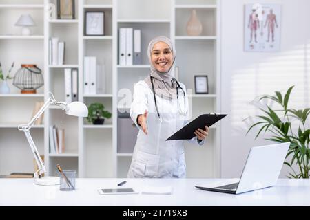 Portrait de femme médecin musulmane heureuse dans le hijab debout à table dans le bureau de l'hôpital, portant un manteau blanc, dossier dans les mains, étendant la main dans l'accueil, souriant et regardant la caméra. Banque D'Images