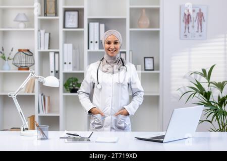 Portrait de femme médecin arabe heureuse dans le hijab debout à table dans le bureau de l'hôpital avec stéthoscope autour du cou, les mains dans les poches, souriant et regardant la caméra. Banque D'Images
