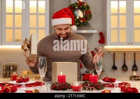 Heureux jeune homme dans le chapeau de Santa ouvrant cadeau de Noël à la table dans la cuisine Banque D'Images