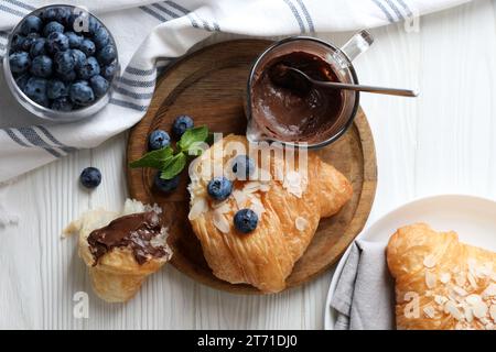 Délicieux croissants aux flocons d'amandes, chocolat et myrtilles servis sur une table en bois blanc, à plat Banque D'Images