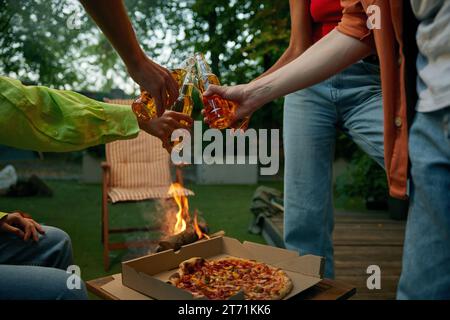 Amis grillage en pique-nique en buvant de la bière et en mangeant de la pizza à proximité du feu de joie Banque D'Images