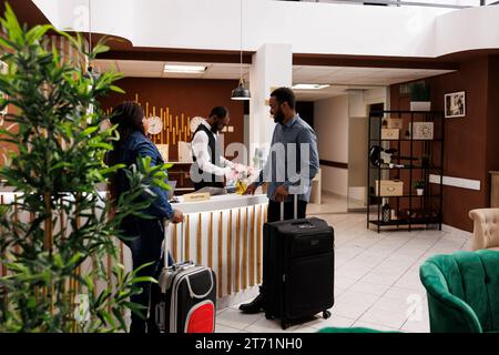 Heureux couple noir debout à la réception de l'hôtel avec des bagages, enregistrement à la station de lune de miel, personnes attendant à la réception pour l'enregistrement. Réceptionniste aidant les clients à régler leur départ Banque D'Images