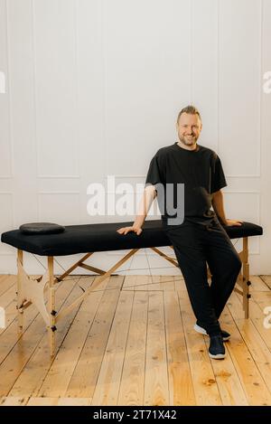Portrait en longueur d'un masseur masculin debout derrière un lit de massage professionnel Banque D'Images