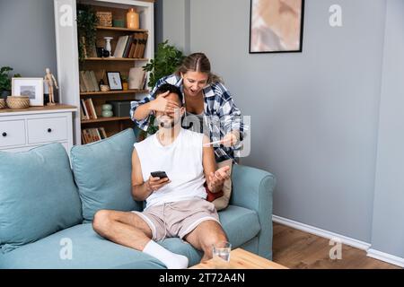 Happy Wife fait la surprise au mari, couvre ses yeux et tient le test de grossesse, informe qu'il va devenir père va partager de bonnes nouvelles, poser ensemble Banque D'Images