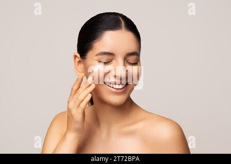 Protection saisonnière de la peau du visage. Photo de beauté de la femme multiraciale magnifique avec le sourire incroyable appliquant de la crème sur son visage Banque D'Images