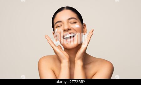 Photo de beauté de la femme multiraciale magnifique avec un sourire incroyable appliquant un hydratant sur son visage. Protection saisonnière de la peau du visage Banque D'Images