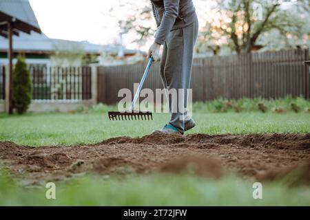 Homme adulte méconnaissable portant un survêtement gris debout sur la pelouse verte et le sol de travail avec râteau de jardin. Humain avec objet agricole dans les mains loos Banque D'Images