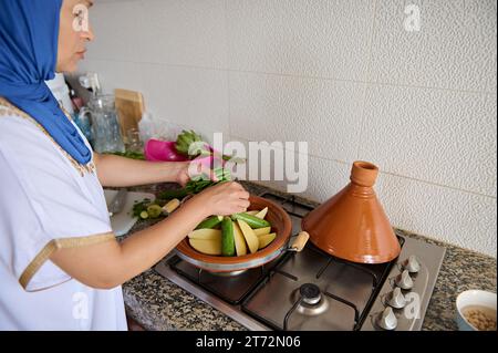 Portrait authentique d'une femme musulmane du Moyen-Orient avec la tête couverte de hijab, mettant des haricots verts frais sur des légumes, fumant dans un pot d'argile tajine. Banque D'Images