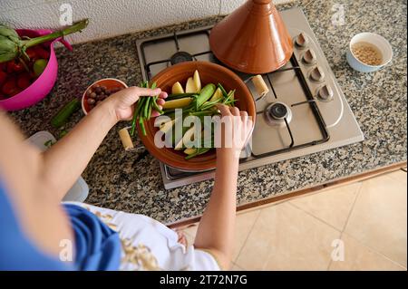 Vue de dessus d'une femme au foyer avec la tête couverte de hijab, mettant des haricots verts frais sur les légumes, fumant dans un pot d'argile tajine. Cuisine marocaine, culi Banque D'Images