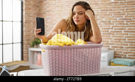 Jeune belle femme hispanique faire selfie par smartphone appuyé sur le panier avec des vêtements à la buanderie Banque D'Images