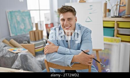 Jeune homme caucasien confiant, un artiste barbu, souriant avec éclat tout en étant assis au cœur de l'atelier d'art animé, pleinement immergé dans la joie de Banque D'Images