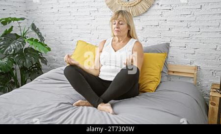 Éveillée et concentrée, femme blonde d'âge moyen aime l'exercice de yoga matinal assis sur le lit, embrassant le calme et l'équilibre dans sa chambre à coucher Banque D'Images