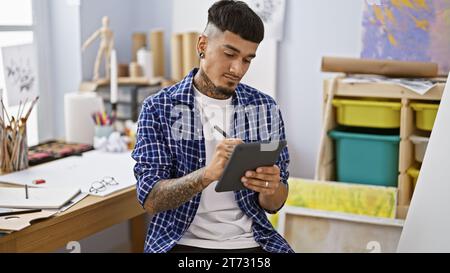 Jeune homme latin détendu, artiste tatoué, sérieusement plongé dans le dessin d’un portrait sur son pavé tactile dans un atelier d’art, incarnant la créativité. Banque D'Images