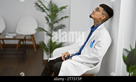 Jeune médecin latin épuisé en manteau blanc, assis stressé dans la salle d'attente de la clinique, son beau portrait gravé de concentration Banque D'Images