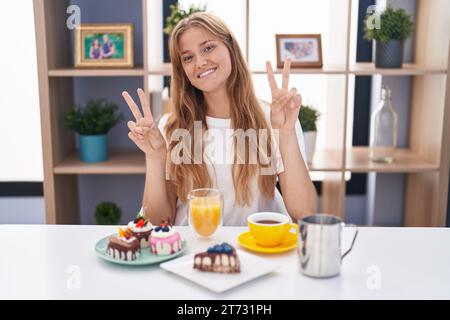 Jeune femme caucasienne mangeant des pâtisseries t pour le petit déjeuner souriant regardant la caméra montrant les doigts faisant signe de victoire. numéro deux. Banque D'Images