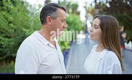 Un père et une fille hispaniques confiants s'engagent dans une conversation chaleureuse, debout ensemble, souriant, rayonnant la positivité au cœur du parc ensoleillé Banque D'Images