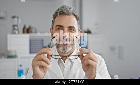 Jeune homme hispanique souriant aux cheveux gris, plein de confiance, tenant des lunettes au laboratoire comme un scientifique joyeux au travail Banque D'Images