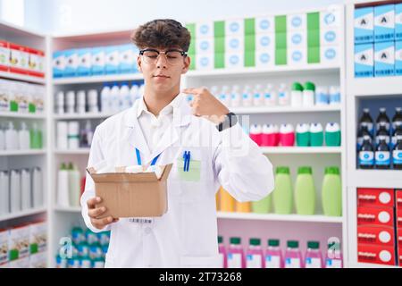 Adolescent hispanique travaillant à la pharmacie pharmacie tenant la boîte avec des pilules pointant du doigt à un auto souriant heureux et fier Banque D'Images