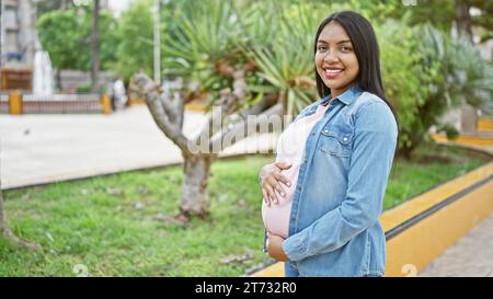 Joyeuse jeune femme latine, attendant joyeusement la maternité, touchant avec confiance son ventre enceinte, souriant dans la lumière du soleil, debout tranquillement au milieu de la ville Banque D'Images