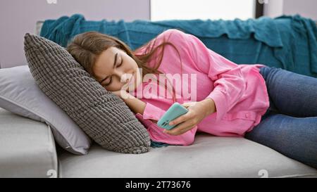 À la maison, portrait d'une belle jeune femme hispanique au visage sérieux concentrée sur l'envoi de SMS, en utilisant son smartphone tout en étant allongée sur le canapé du salon Banque D'Images