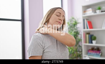 Portrait d'une jeune femme blonde en détresse, attrayante, souffrant d'un mal de cou sévère, debout malheureusement à l'intérieur dans sa maison confortable Banque D'Images