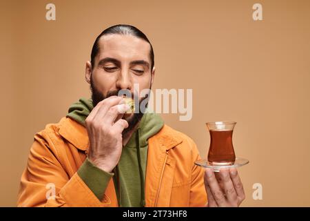 homme barbu heureux tenant le thé turc dans une tasse en verre traditionnelle et mangeant de savoureux baklava sur beige Banque D'Images