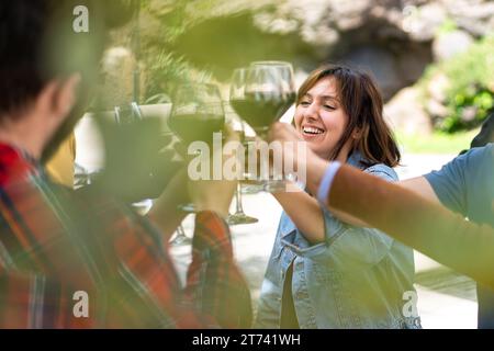 Un moment de franchise capturé alors que des amis toastent avec des verres de vin rouge, partageant le bonheur dans un cadre extérieur, avec l'accent sur une femme souriante. Banque D'Images